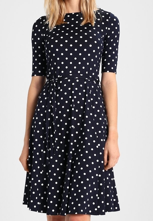 Femme portant une robe bleu marine longueur genou avec des pois blancs, manches aux coudes et ceinture nouée à la taille.