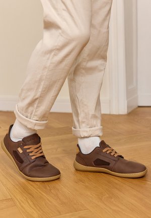 Person trägt beigefarbene Hose, weiße Socken und braune legere Lederschuhe und steht auf einem Holzboden im Innenraum.