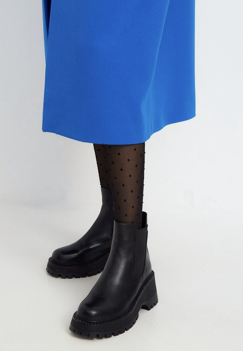Bottines noires à talons épais portées avec des collants noirs transparents à pois et une jupe midi bleu vif sur fond blanc.