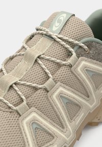 Shoe atlético de um tom claro de bege com parte superior em malha, detalhes texturizados, atacadores elásticos e sola de borracha. Logótipo notável na lingueta.