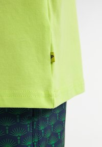 Limoengroene katoenen T-shirt met een rechte zoom en zijlabel; gecombineerd met marineblauwe shorts met een groen palmenbladpatroon.