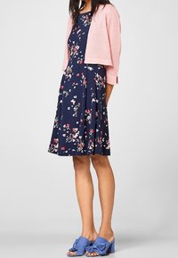 Květinové tmavě modré šaty s rozšířenou sukní, doplněné světle růžovým krátkým svetříkem. Modré nazouvací boty s uzlovým detailem.