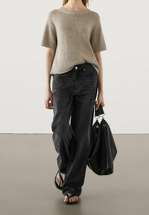 Woman wearing beige short-sleeve knit top, loose black jeans, flip-flops, and carrying a large black tote bag in her right hand.