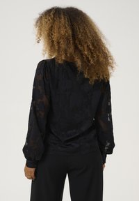 Blusa nera floreale con maniche lunghe trasparenti, tessuto testurizzato e polsini con bottoni. Silhouette morbida con dettagli intricati del motivo floreale.