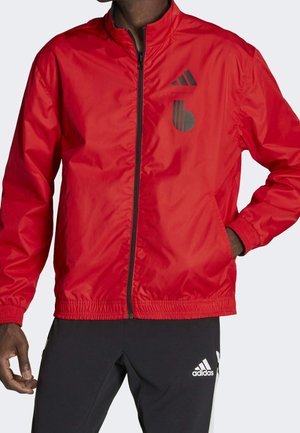 Giacca a vento rossa con zip frontale, polsini ed orlo elasticizzati. Presenta il logo Adidas e accenti grafici in nero. Materiale liscio e leggero.