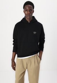 Sort bomulds-hoodie med en lomme foran og et lille logo på venstre bryst, båret over en hvid skjorte og parret med beige bukser.