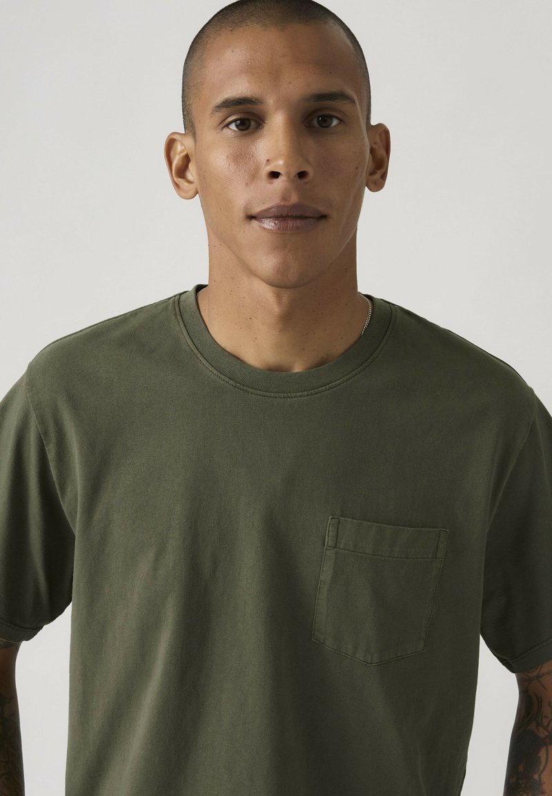 Man met kortgeschoren haar, gekleed in een olijfgroen T-shirt met borstzak, kijkend naar voren tegen een effen lichte achtergrond.