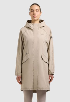 Frau trägt einen knie- bis wadenlangen beigen Kapuzenmantel mit zwei vorderen Pattentaschen, steht vor einem einfarbigen hellen Hintergrund.