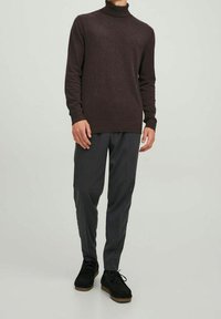 Pull à col roulé marron foncé en matériau doux, associé à un pantalon gris clair fuselé. Chaussures noires à semelle texturée.