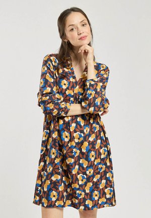 Jeune femme posant avec la main sur le menton, portant une robe longue aux manches longues et aux motifs floraux bleus, jaunes et marron sur fond blanc.