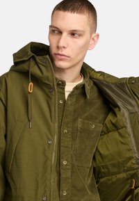 Olivgrön jacka i mjukt tyg, med dragkedja fram, justerbar huva och en skjorta i cordflotta med ficka på insidan.