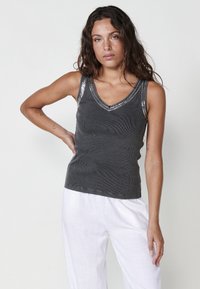 Grijze tanktop met een v-hals, gekenmerkt door een textuurstof en met paillettenaccenten op de schouders. Combinatie met witte, losse broek.