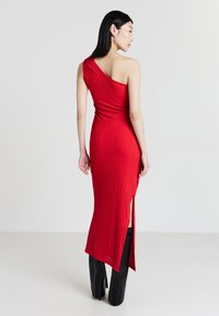 Rotes, geripptes Ein-Schulter-Kleid mit einer figurbetonten Silhouette, das auf einer Seite einen hohen Schlitz hat und eine glatte Textur aufweist. Getragen mit schwarzen, weit geschnittenen Hosen.