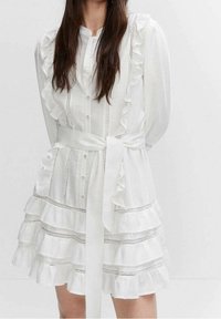 Robe blanche avec manches volantées et ourlet à volants. Elle présente un devant boutonné, une ceinture en tissu et de délicats accents argentés le long des coutures.