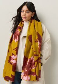 Floralsete mustriga sall sinepikollases toonis, millel on roosad ja burgundia lillemotiivid. Pehme tekstuuriga, drapeeritud disainiga, kantud kerge kampsuni peale.