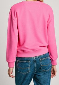 Osoba oblečená v jasně růžovém svetru a modrých džínách, stojící zády k objektivu na jednolitém pozadí.