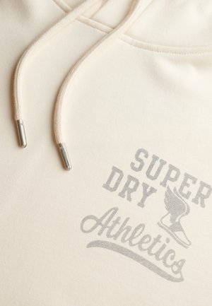 Sweat à capuche couleur crème avec une texture douce, présentant une impression graphique grise indiquant "SUPER DRY Athletics" et un cordon de serrage ajustable avec embouts métalliques.