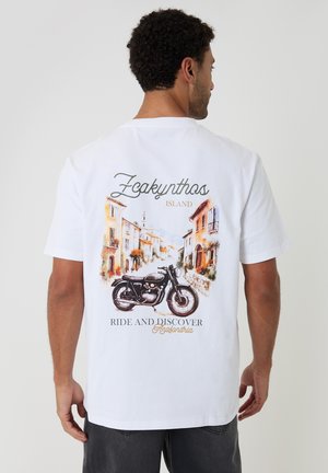 Mand iført hvid T-shirt med farverigt gadebillede fra Zakynthos-øen og klassisk motorcykel grafik, tekst "Kør og opdag."