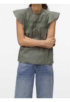 Person iført en grøn bluse med flæser på ærmerne og blonde-detaljer, kombineret med lyseblå jeans, armene krydset over brystet.