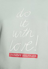 Texto bordado en blanco "¡hazlo con amor!" sobre el logo rojo "TOMMY HILFIGER" en una tela verde clara, probablemente de una sudadera o camiseta.