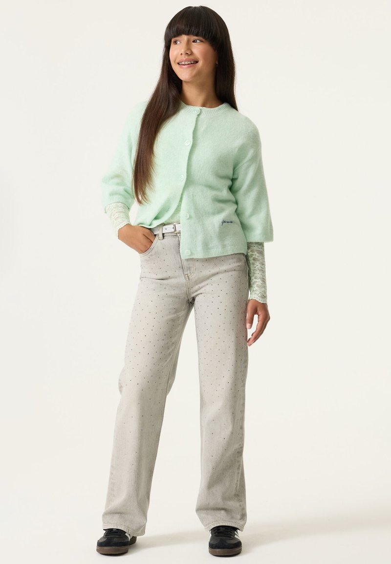 Cárdigan de manga corta verde menta con botones, combinado con jeans gris claro con estampado de lunares y calzado negro.