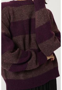 Personne portant un pull douillet rayé large violet et marron à manches longues, ajustant la manche près du visage.