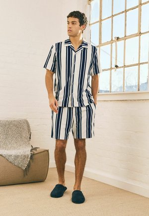 Ung mand, der står indendørs iført en skjorte med lodrette navy og hvide striber og matchende shorts med navy tøfler.