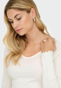 Blondynka z falującymi włosami nosi srebrne kolczyki w kształcie kół, srebrny pierścionek oraz biały prążkowany top z długim rękawem i falbaniastymi mankietami.