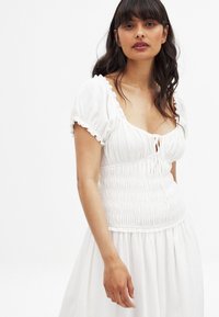 Vit klänning med en figurnära, volangförsedd överdel, smockdetaljer och puffärmar. Kjolen flödar mjukt och har ett lätt, texturerat tyg.