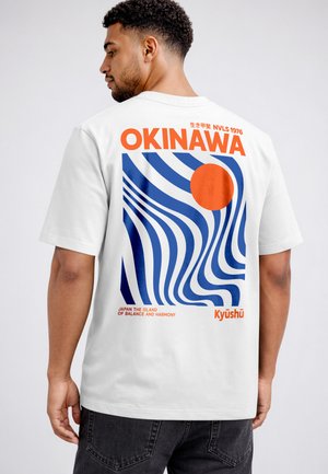 Man wearing white T-shirt with bold orange "OKINAWA" text and blue wavy lines design on back, paired with black jeans.