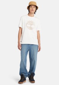 T-shirt en coton blanc avec un graphique d'arbre circulaire en beige, associé à un jean bleu à coupe ample et des bottes marron avec des accents noirs.