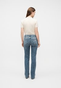 Γυναίκα που στ έκεται με την πλάτη προς τα εμπρός, φοράει κρεμ μπλούζα με κοντά μανίκια και ψηλόμεσο μπλε τζιν με πίσω τσέπες και μικρό λογότυπο.