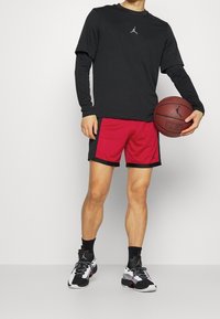 Persona con una camiseta de manga larga negra, pantalones cortos de baloncesto rojos y negros, calcetines negros y zapatillas blancas y negras, sosteniendo un balón de baloncesto marrón.