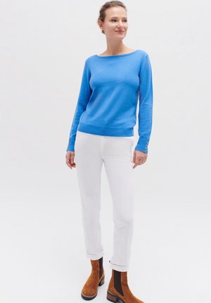 Blauwe lichtgewicht trui met een brede halslijn, een getailleerde taille en knoopaccenten op de mouwen. Gecombineerd met witte broek en bruine enkellaarsjes.