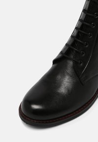 Botín de cuero negro hasta el tobillo con una textura suave, punta redondeada y cinco pares de ojetes para los cordones. Presenta un detalle de costura en rojo contrastante.