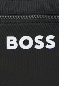Sac en tissu noir doté d'une surface texturée avec un logo "BOSS" blanc bien en évidence. Comprend une fermeture éclair en haut.