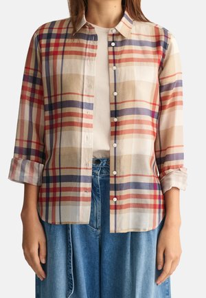 Frau trägt ein knöpfbares, langärmeliges kariertes Hemd mit beigen, roten und blauen Streifen über einem weißen Top und hoch taillierten blauen Jeans.