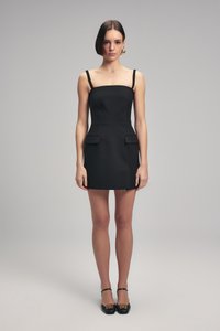 Schwarzes, figurbetontes Kleid mit einem quadratischen Ausschnitt, dünnen Trägern und zwei vorderen Taschen. Das Material wirkt glatt und strukturiert. Dazu wurden schwarze Schuhe getragen.