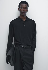 Zwarte polo met lange mouwen, een kraag en knoopsluiting, gecombineerd met op maat gemaakte zwarte broek en een zwarte leren tas.