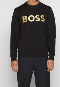 En person som bär en svart sweatshirt med en stor guld "BOSS"-logotyp står mot en neutral bakgrund och visar plaggstil och passform.