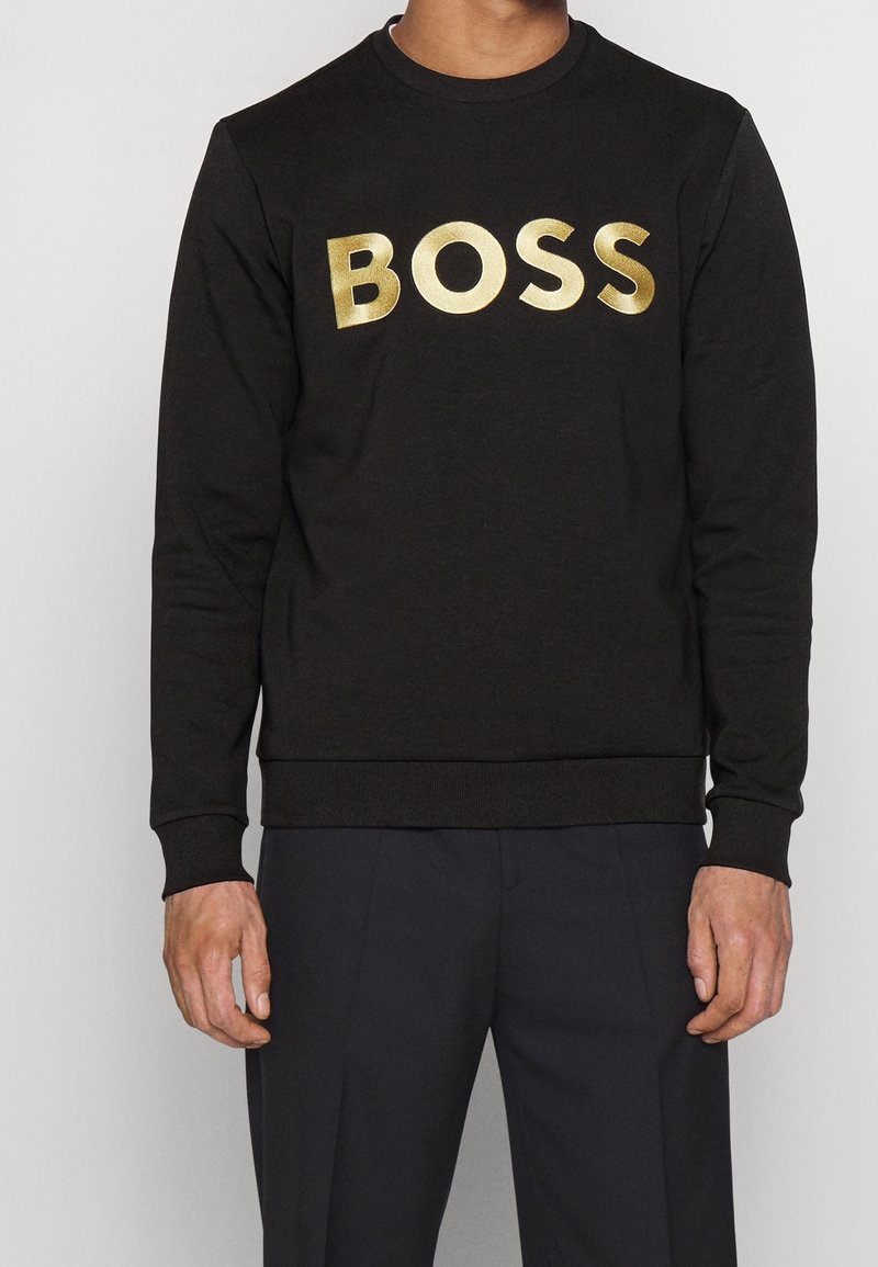 En person som bär en svart sweatshirt med en stor guld "BOSS"-logotyp står mot en neutral bakgrund och visar plaggstil och passform.