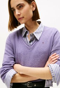 Suéter de pico lavanda sobre una camisa de cuello azul claro y blanca con rayas, con mangas enrolladas y un pequeño logo en el suéter.