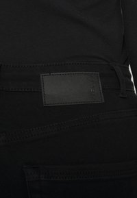 Černé džíny s hladkou texturou, které mají jednoduchou černou koženou nášivku na pase a jemné detaily v prošívání.