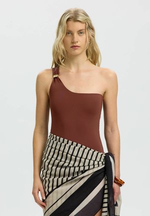 Donna con lunghi capelli biondi che indossa un top marrone monospalla e una gonna avvolgente a righe bianco e nero su uno sfondo semplice.