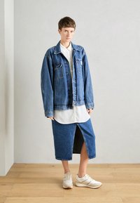 Jeansjacke mit großen Taschen und Knöpfen, über einem weißen Hemd getragen. Das Model trägt einen Jeansrock mit einem Schlitz an der Vorderseite und weiße Sneaker.