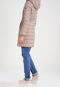 Ljust beige pufferjacka med huva, med horisontellt quiltat mönster och sidofickor. Bärs med blå jeans och vita sneakers.