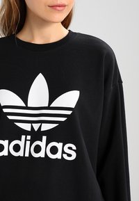Kvinna som bär en svart sweatshirt med en stor vit Adidas trefoil-logotyp och varumärkesnamn på framsidan, avskuren vid axlarna.