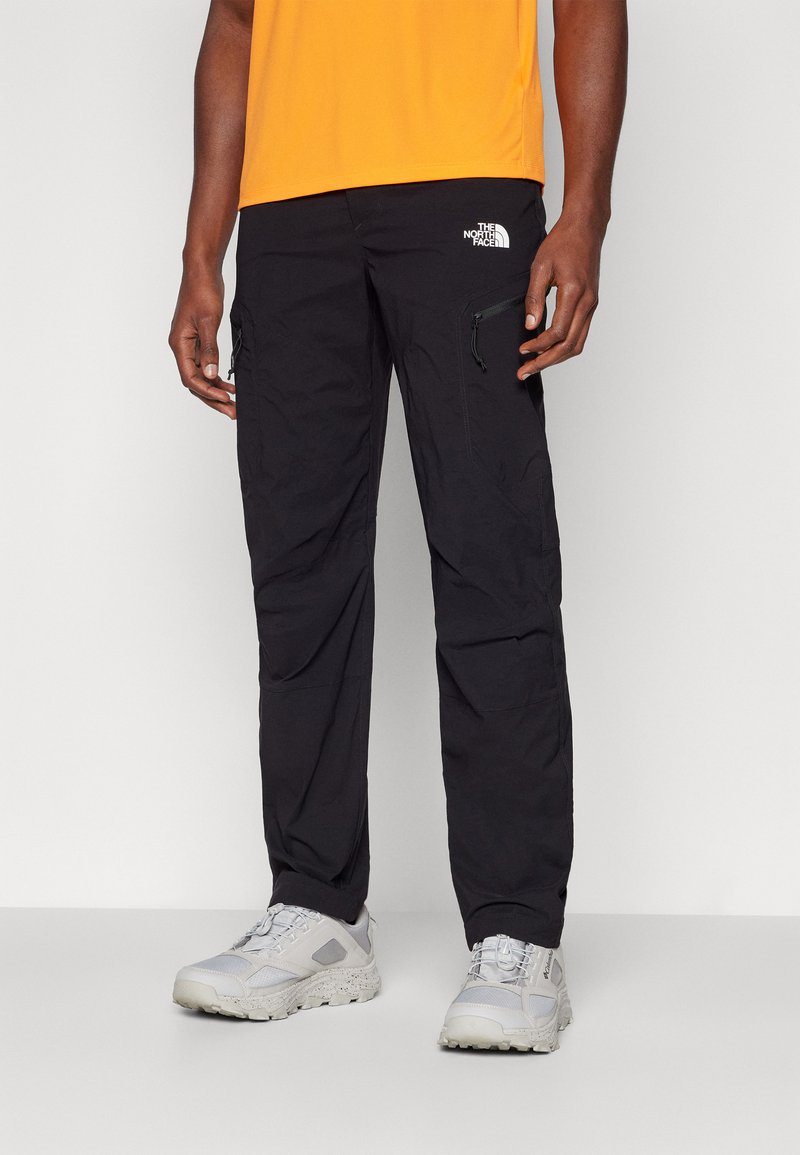 Crne vanjske hlače s džepovima na patentni zatvarač, podesivim manžetama i logotipom The North Face. Nosive s svjetložutim cipelama i narančastom majicom.