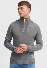 Szary dzianinowy sweter z wysokim kołnierzem i połową zamka, z prążkowaną teksturą oraz małą łatką z logo w lewym dolnym rogu.