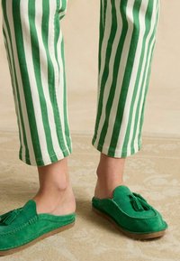 Zelené semišové topánky na jemné nazutie s detailom strapcov, v kombinácii s pruhovanými nohavicami v zelenej a bielej farbe. Tkanina je mäkká s kontrastnými živými farbami.